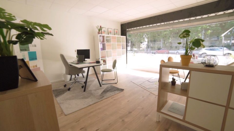 Bureau moderne avec ordinateur, fauteuil ergonomique, plante verte et étagère en bois dans un espace de coworking lumineux.