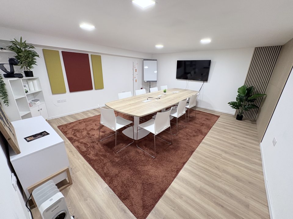 Salle de réunion en sous-sol avec table en bois, chaises blanches, écran plat et paperboard.